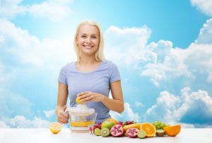 healthy eating, vegetarian food, diet, detox and people concept - smiling woman with squeezer squeezing fruit juice over blue sky and clouds background